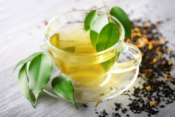 Glass cup of tea with green leaves and scattered tea around on grey wooden background