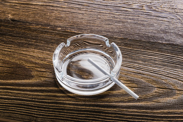 Empty glass ashtray on a wooden table and thin white cigarette.