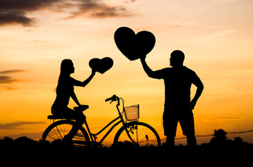 Naklejka premium Silhouette of a man and a woman riding a bicycle together and holding big hearts