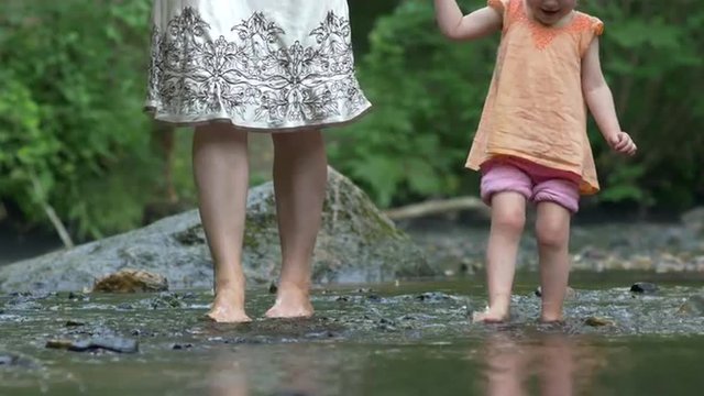 Mother and a young daughter paddle barefoot over rocks in a shallow creek