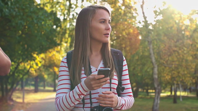 Young woman walking with camera in the park and texting a message on her smartphone