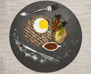 Big juicy grilled steak marbled beef with egg baked potatoes with barbecue sauce. Served on a stone plate with a fork and spoon on a textile background.