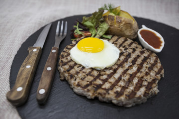 Big juicy grilled steak marbled beef with egg baked potatoes with barbecue sauce. Served on a stone plate with a fork and spoon on a textile background.