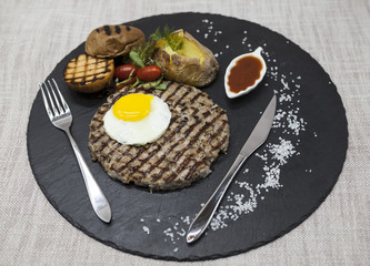 Big juicy grilled steak marbled beef with egg baked potatoes with barbecue sauce. Served on a stone plate with a fork and spoon on a textile background.