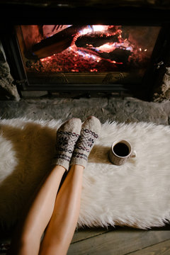 Feet in woollen socks by the Christmas fireplace. Woman relaxes by warm fire with a cup of hot drink and warming up her feet in woollen socks. Close up on feet. Winter and Christmas holidays concept