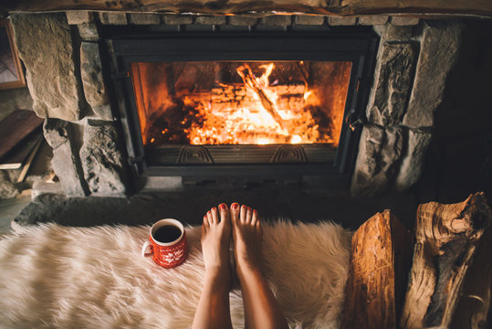 Bare woman feet by the cozy fireplace. Woman relaxes by warm fire with a cup of hot drink and warming up her feet. Close up on feet. Winter and Christmas holidays concept
