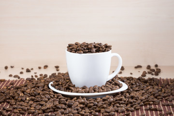 White full of roasted coffee beans cup standing on a white plate on tablemat on light wooden textured background.