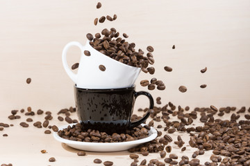 Roasted coffee beans falling down into white cup standing on a black cup on white plate standing on light wooden textured background.