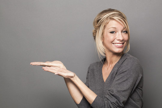 body language concept - smiling 20s blond woman showing an empty display of a product for an advertisement,studio shot on gray background