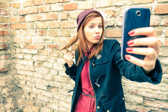Young pretty woman taking selfie outdoors - Female winter fashion portrait - Teenager student holding mobile phone for selfi photo next to brick wall background - Soft and hazy vintage filtered look 