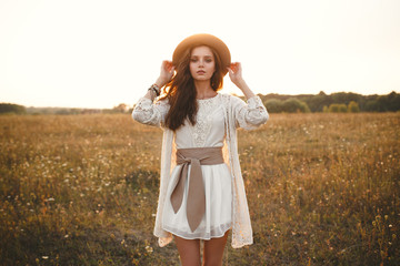 Fashion portrait of beautiful young pretty girl with hippie outfit holding hat outdoors at sunset....