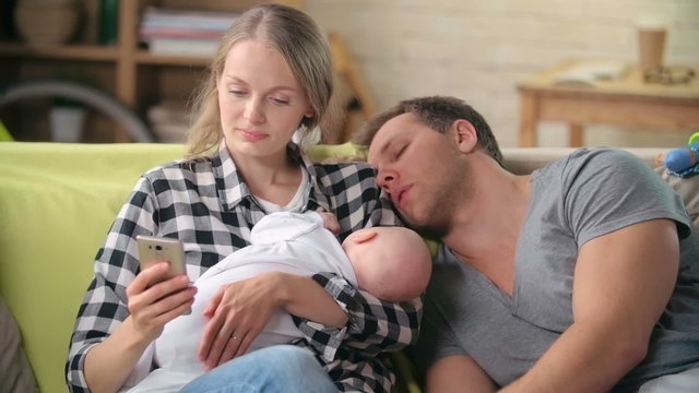 Tired young mother text messaging on a smart phone, her baby sleeping in her arms and her husband napping on her shoulder