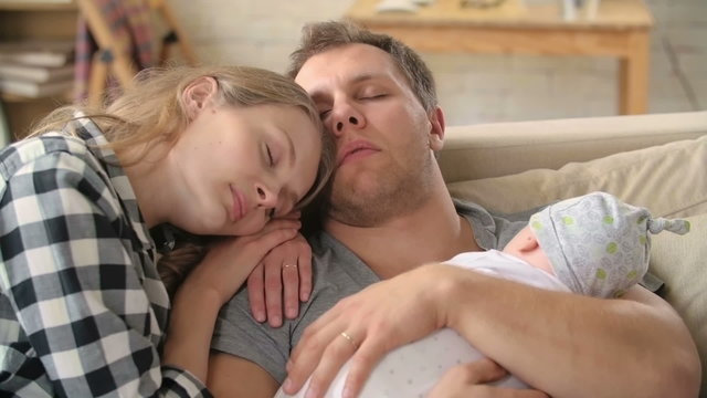 Tired young parents sitting and sleeping on sofa with their newborn baby 