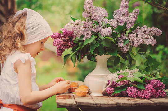 Cute Child Girl On Cozy Outdoor Tea Party In Spring Garden With Bouquet Of Lilacs