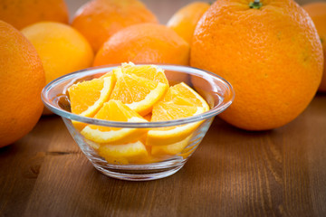 Couple of orange slices in a glass bowl, on a wooden surface with whole oranges in the background