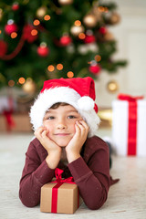 Merry Christmas! The boy with a gift in santa hat lying under the Christmas tree.