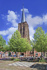 Village square with ancient brick church, green trees, red pavers, blue sky and clouds.