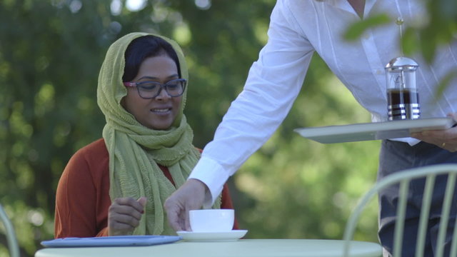 Woman receiving a coffee whilst sitting at a table outside using a digital tablet
