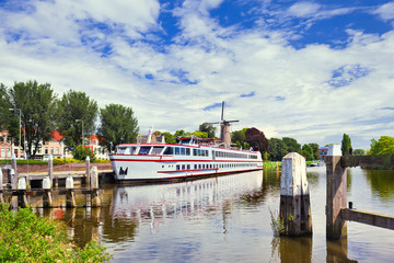 Fototapeta na wymiar White tour-boat moored in a tranquil canal with a windmill on the background, Gouda, The Netherlands