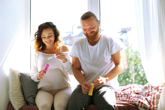 Happy beloved couple blow soap bubbles in waiting for baby's birth on window board