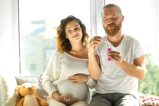 Happy beloved man and pregnant woman blow soap bubbles in waiting for baby's birth on window board