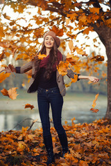 slender young brunette beautiful girl with long hair in a beret with a scarf and coat walks in autumn park with a lot of yellow and orange leaves