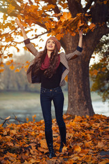 slender young brunette beautiful girl with long hair in a beret with a scarf and coat walks in autumn park with a lot of yellow and orange leaves