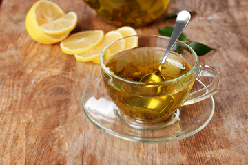 Glass cup of tea and teapot on wooden background decorated with sliced lemon and green leaves, close up