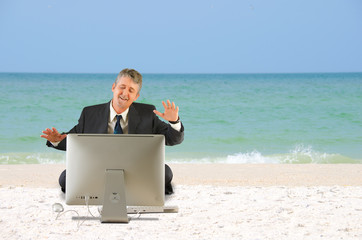 A business man in a suit is sitting on the beach with a big computer in front of him and he has a...