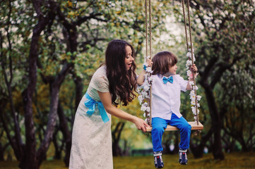 Naklejka na ściany i meble Happy loving mother and toddler son on swing in spring garden