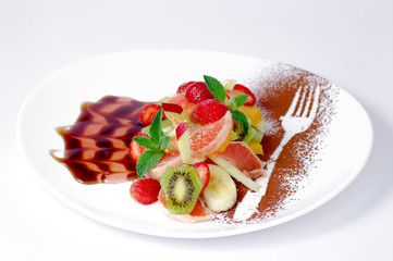 close-up of fruit salad, decorated with mint leaces, cinnamon powder and brown sauce, on oval white plate, on a white background