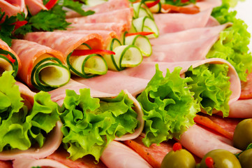 descending close-up of rolls of ham and salami decorated with green olives, leaves of lettuce, slices of cucumber and kiwi and leaves of parsley