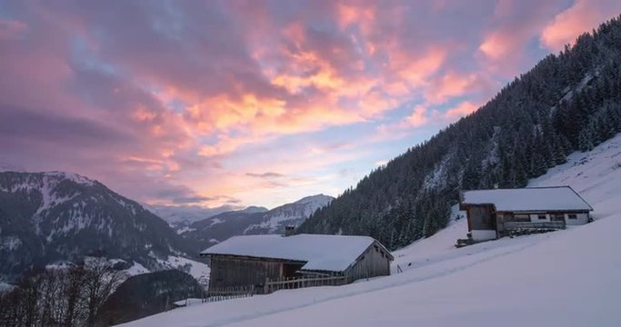 Time lapse of an epic winter sunset over a valley in Austrian Alps with a shed and house covered with snow in foreground