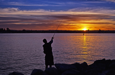 Silhouette of fisherman on the quiet bay with the rays of sunset