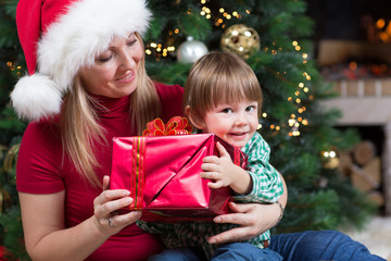 Happy woman gives wrapped christmas presents gifts to child baby toddler sitting near Christmas tree