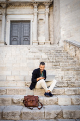 Young businessman sitting on steps of city building and using his phone