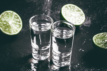 Vodka shot glass on black wet surface with limes.