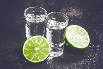 Vodka shot glass on black wet surface with limes.