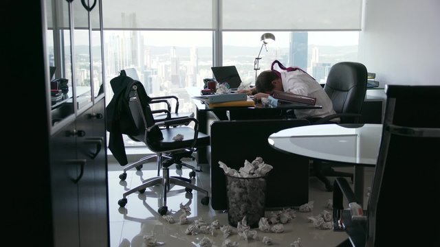 Corporate manager in modern office tries to write a job letter. The man is frustrated and keeps on screwing up paper. He bangs his head against the table holding his tie. Wide angle shot