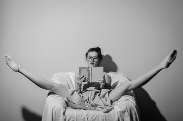 Black and white photography of beautiful young lady wearing glasses and holding a book relaxing split legs in chair