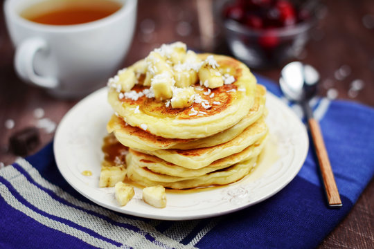 Breakfast: cup of green tea and stack of pancakes with banana slices and coconut flakes on vintage plate on dark blue kitchen towel