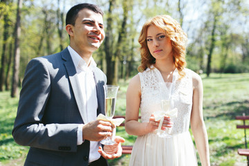 blonde stylish bride in white dress and happy groom drinking wedding champagne in park