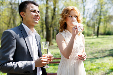 blonde stylish bride in white dress and happy groom drinking wedding champagne in park