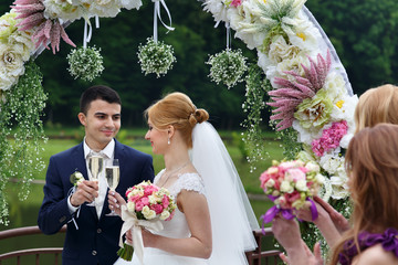 Stylish blonde bride with flowers and handsome groom under wedding arc drinking champagne