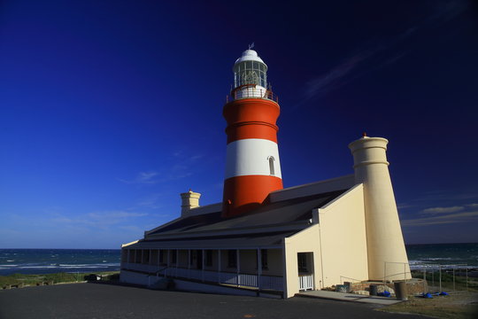 Cape L'Agulhas lighthouse / A lighthouse painted in red and white with a building at the bottom and a deep blue and clear sky and sea in the background.