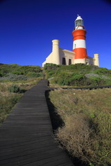 Fototapeta na wymiar Cape L'Agulhas lighthouse (II) / A lighthouse painted in red and white with a building at the bottom and a wooden boardwalk leading to it. Deep blue and clear sky in the background.