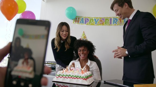 Business woman celebrating her birthday and doing a party with colleagues in her office. A friend records a video on mobile phone of her blowing out clandles on cake. Medium shot