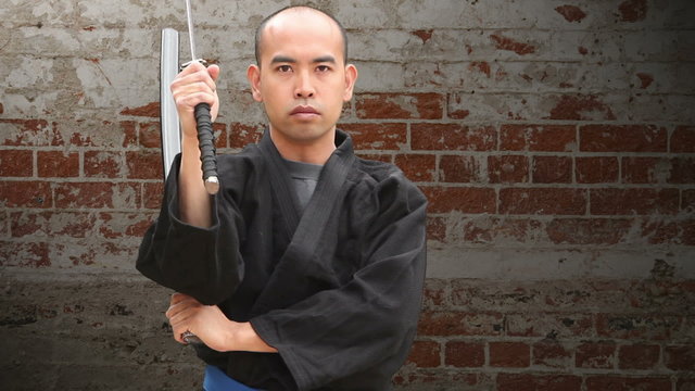 Asian man in a Japanese Gi draws a Samurai Sword, and slashes through the air with sword and scabbard (siniwali style), then resheaths his blade in front of a brick background, with pan and zoom.