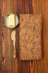 Ржаной домашний хлеб с кунжутом рядом с куском сливочного масла на блюдце и деревянным ножом на деревянном столе