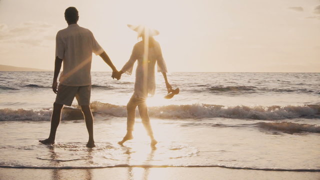 Sunset Walk On A Luxury Beach. Older Couple Holds Hands And Walks Down The Beach At Sunset Getting Their Feet Wet